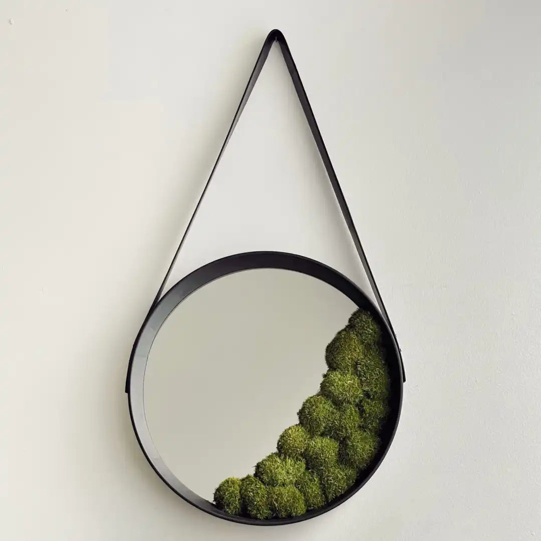Espejo circular con borde negro y decoración de musgo preservado en la parte inferior derecha, colgado en la pared con una correa de cuero.