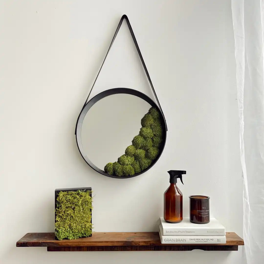 Espejo circular con borde negro y decoración de musgo preservado en la parte inferior derecha, colgado en la pared con una correa de cuero.