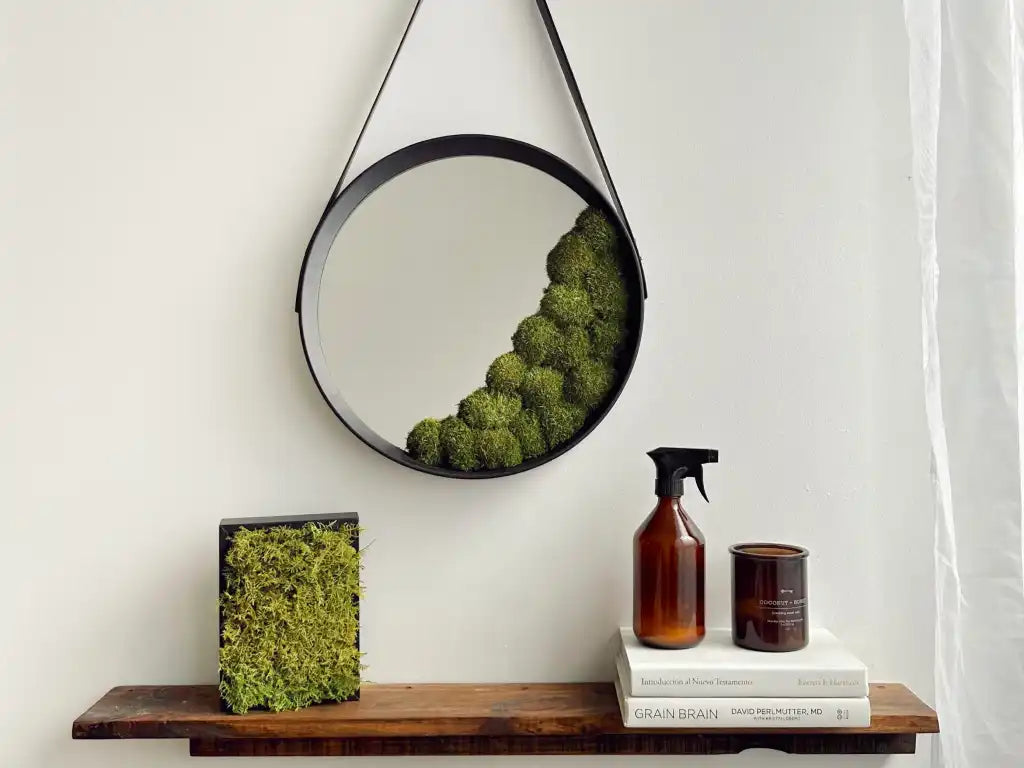 Espejo circular con borde negro y decoración de musgo preservado en la parte inferior derecha, colgado en la pared con una correa de cuero, acompañado de un cuadro de musgo y una botella rociadora sobre una estantería.