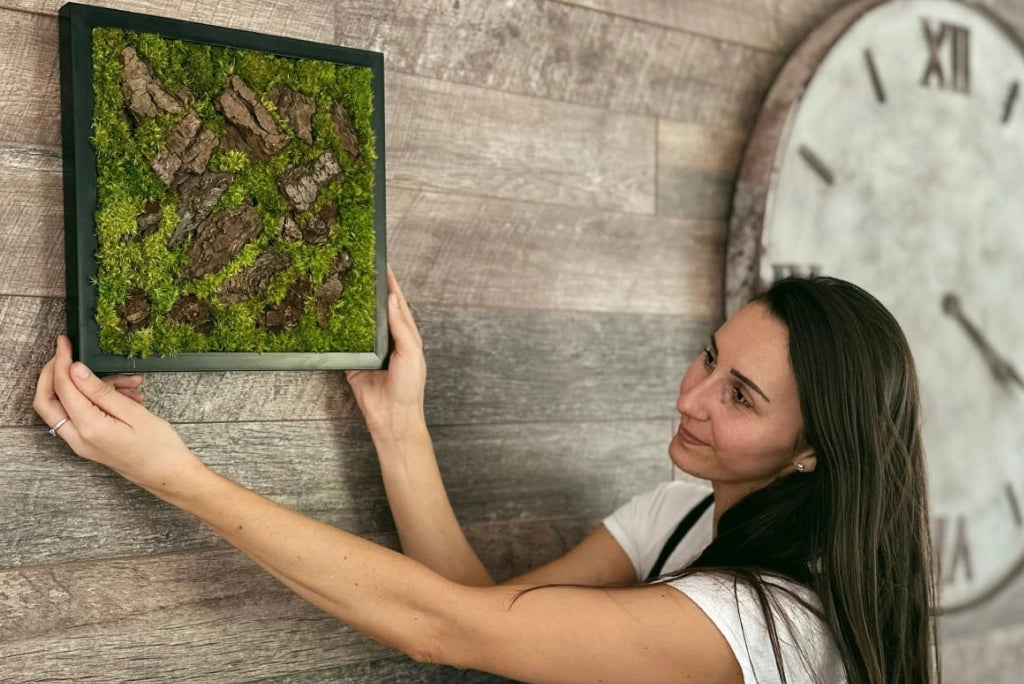 Mujer colgando un cuadro de musgo preservado y corteza en una pared de madera, mostrando el proceso de decoración.